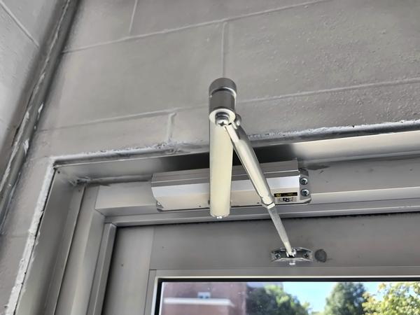 Overhead door closer and arm mounted on the header of an aluminum entrance door, with campus buildings visible outside