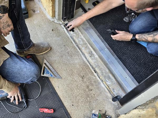 Technicians working at the base of an entrance door, fitting a new aluminum threshold with tools on the mat