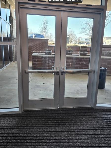 Pair of aluminum-and-glass exterior entrance doors with push bars, seen from inside over a walk-off mat