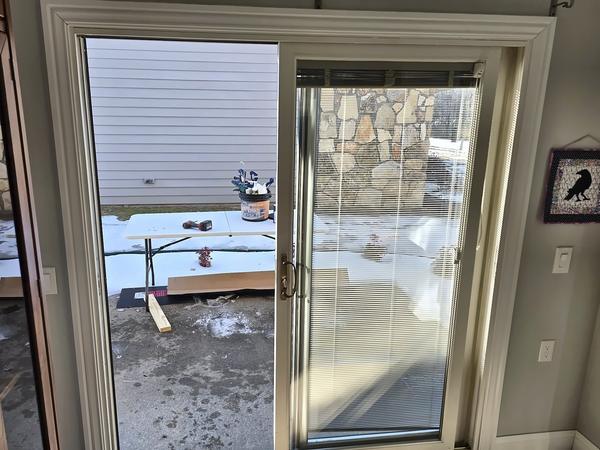 Interior view of an older beige patio sliding door with blinds, looking out to a snowy patio