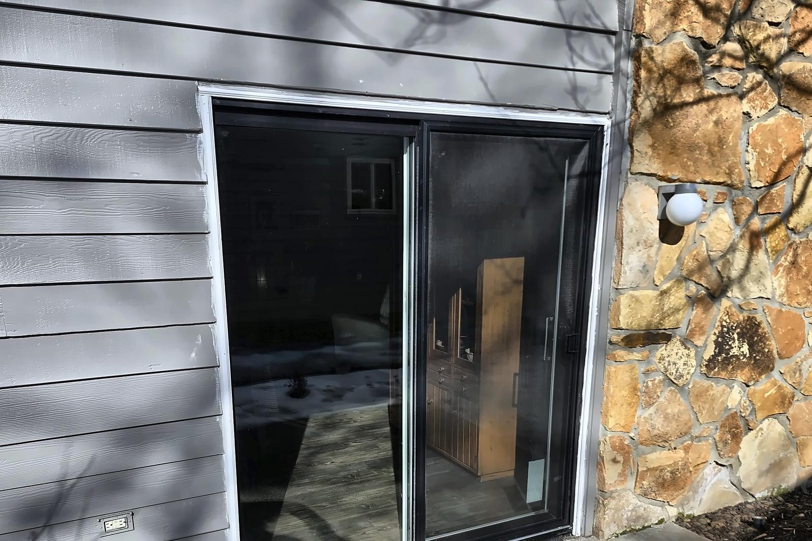 New black-framed patio sliding door installed in gray siding next to a stone wall